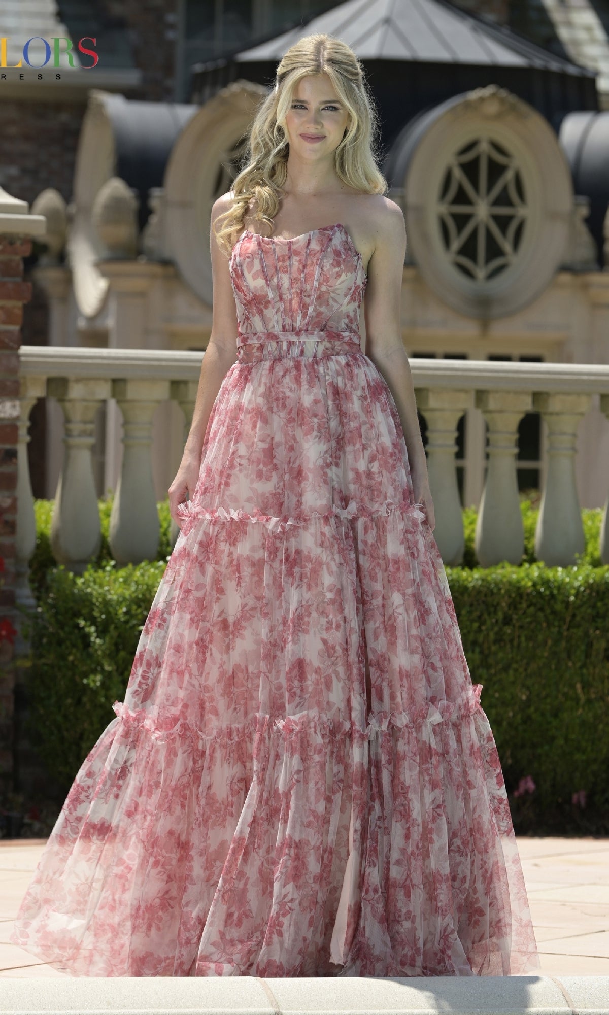 A woman smiles outdoors in the Strapless Long Print Prom Dress: Colors Dress 3810, her long blonde hair flowing. Architectural accents and greenery create a beautiful backdrop for this elegant evening gown.